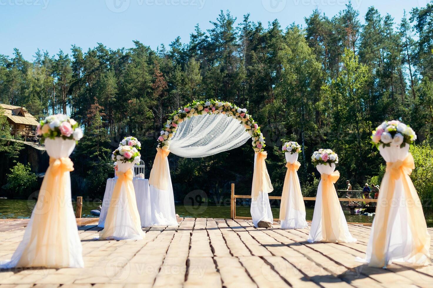 Beautiful place for wedding ceremony on the wooden bridge over the river. Archway decorated with creamy and white cloth and flowers on the natural forest background in summer. photo