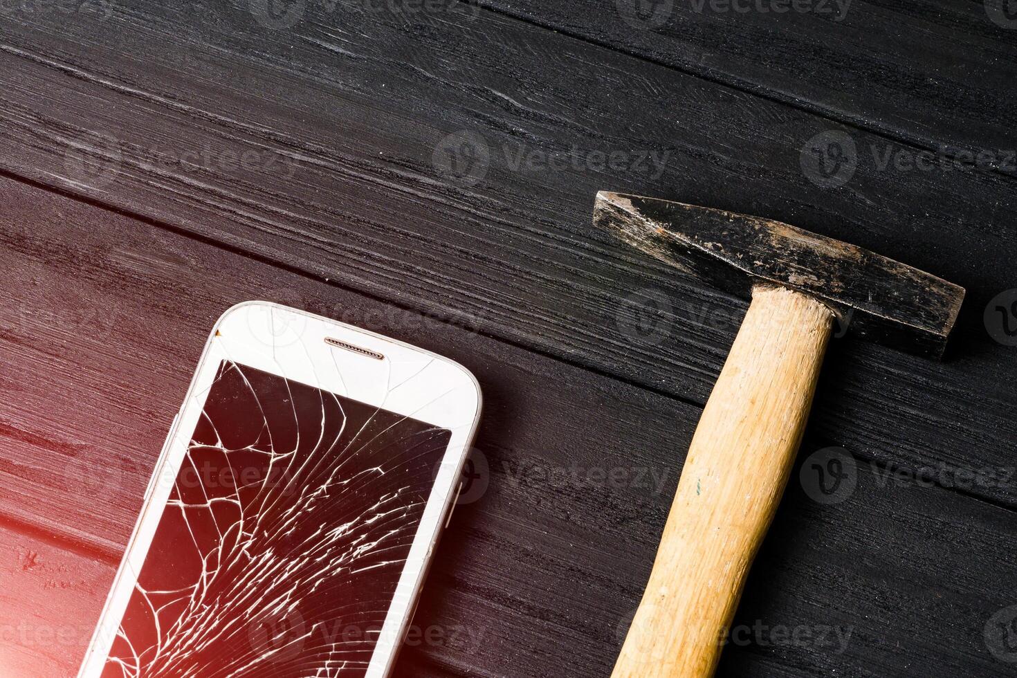 Composition of a hammer and a smart phone with a broken screen over the dark wooden surface. The concept of a device failure photo