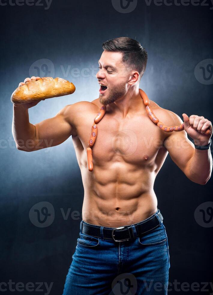 Pleased muscular guy with beautiful naked torso holding a loaf of bread and sausages on his neck. Ready to bite. Harmful food. Lifestyle concept photo