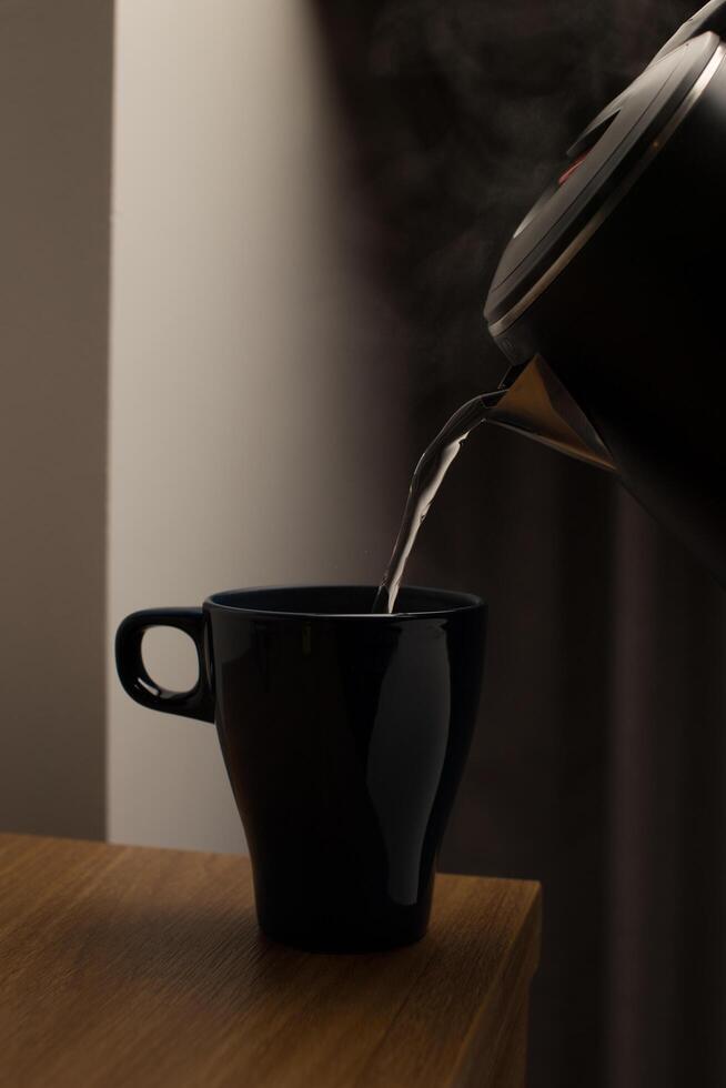 Pour hot water from an electric kettle into a dark blue ceramic cup on a wooden table in the middle of the night. photo