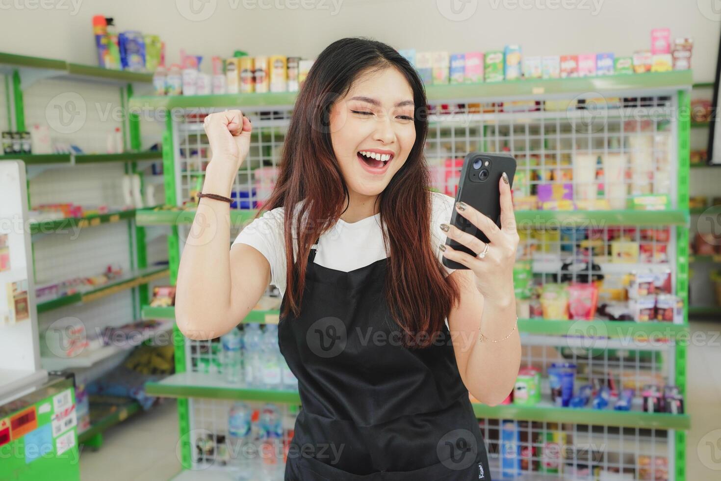 emocionado asiático mujer como un cajero es vistiendo negro delantal y participación su teléfono pantalla foto
