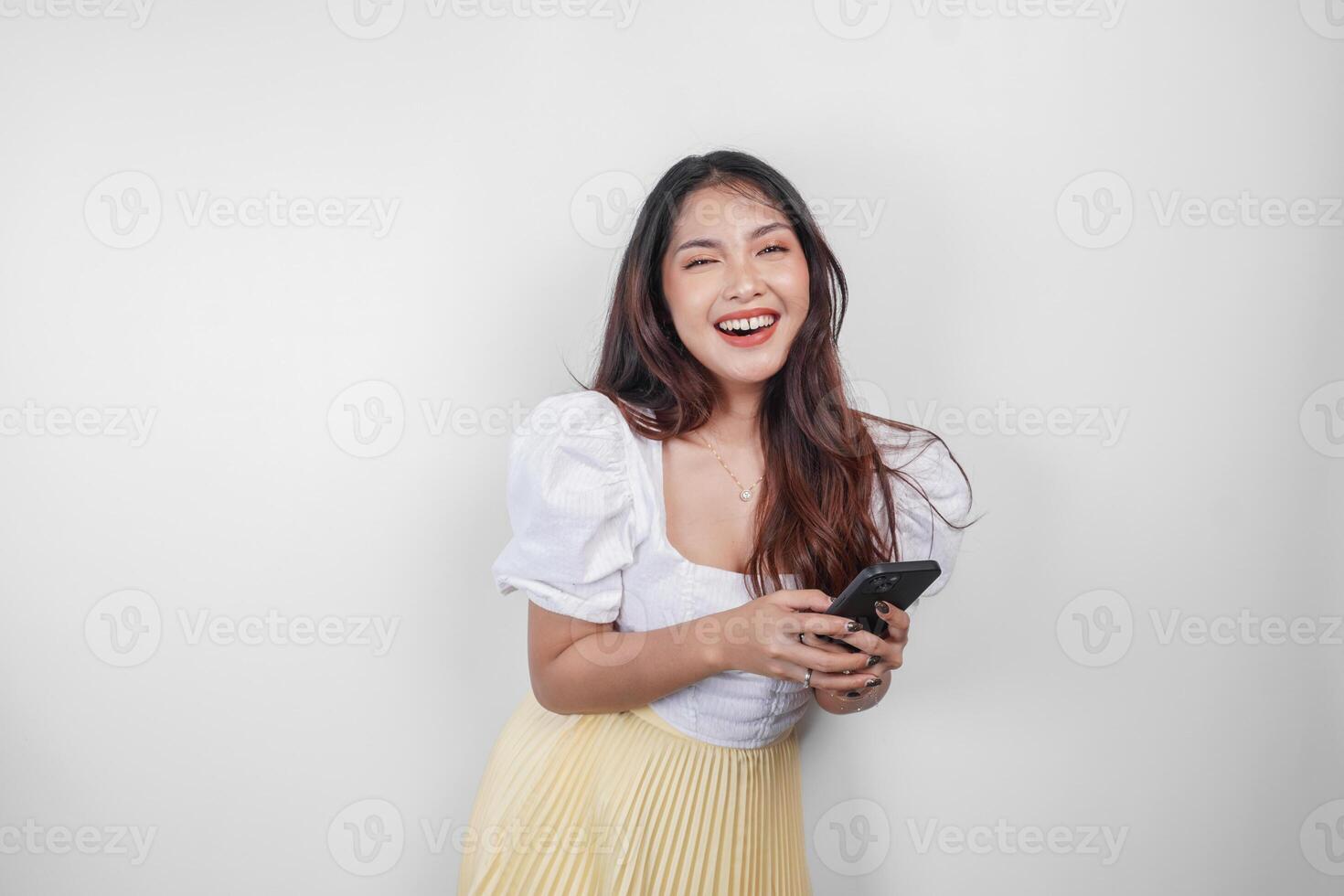 A cheerful Asian woman is smiling and holding her smartphone, isolated by white background. photo