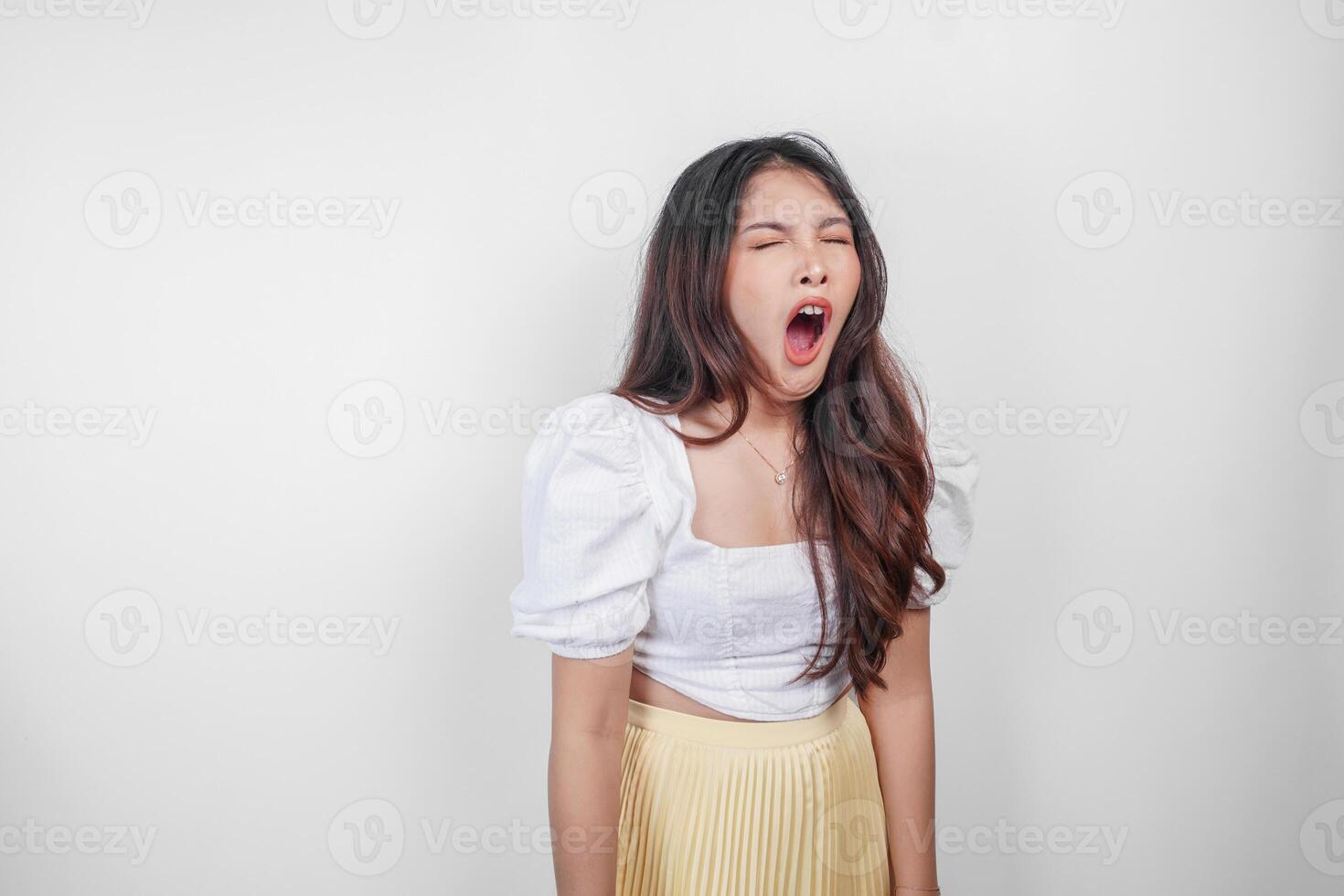 Portrait of sleepy attractive Asian woman wearing a dress, feeling tired after night without sleep, yawning with her mouth wide open photo