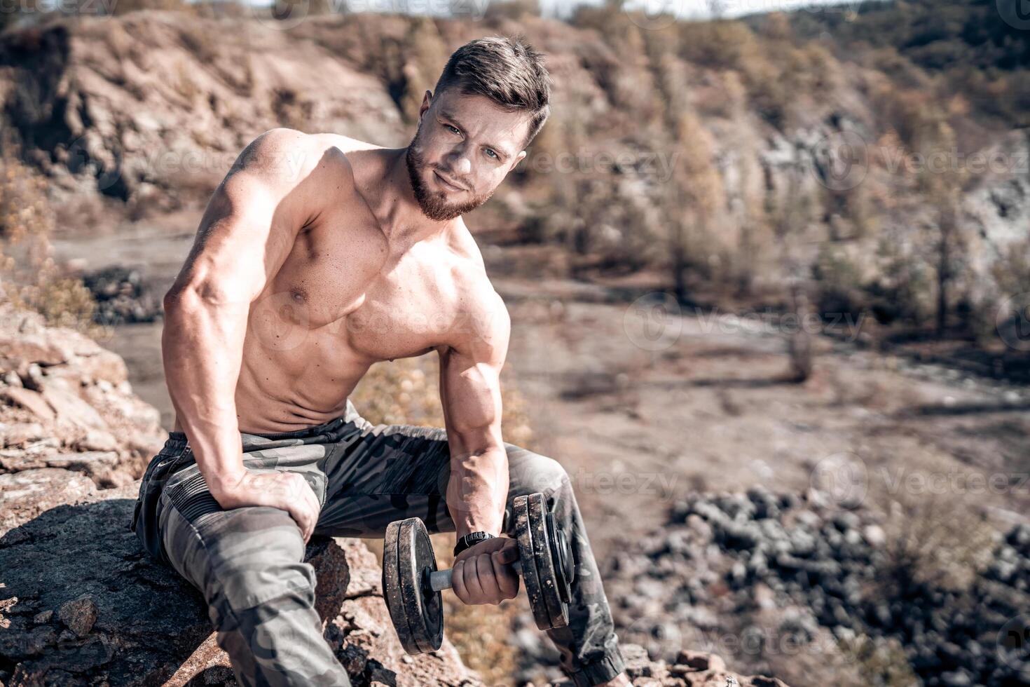 Attractive Caucasian man pumping up muscules sitting on a rock. Muscular athlete holding a heavy dumbbell with one hand. Bodybuilder with naked torso doing exercise with a dumbbell. photo