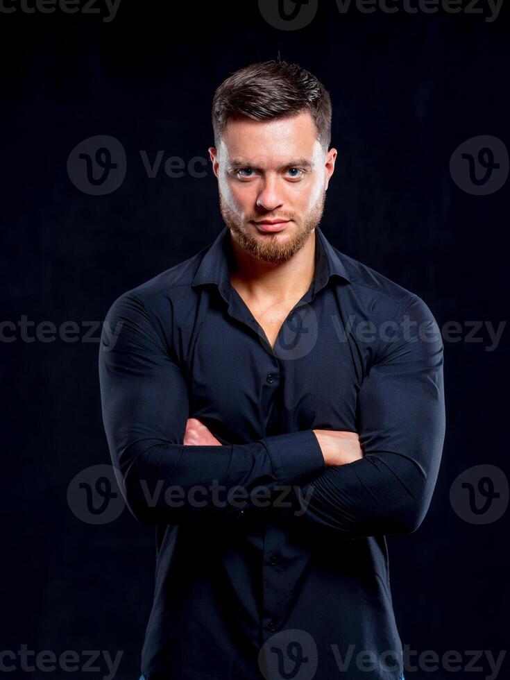 Young man in black shirt smiling isolated on dark background. Standing cross hands. Muscular male, athletic. Fashion portrait photo
