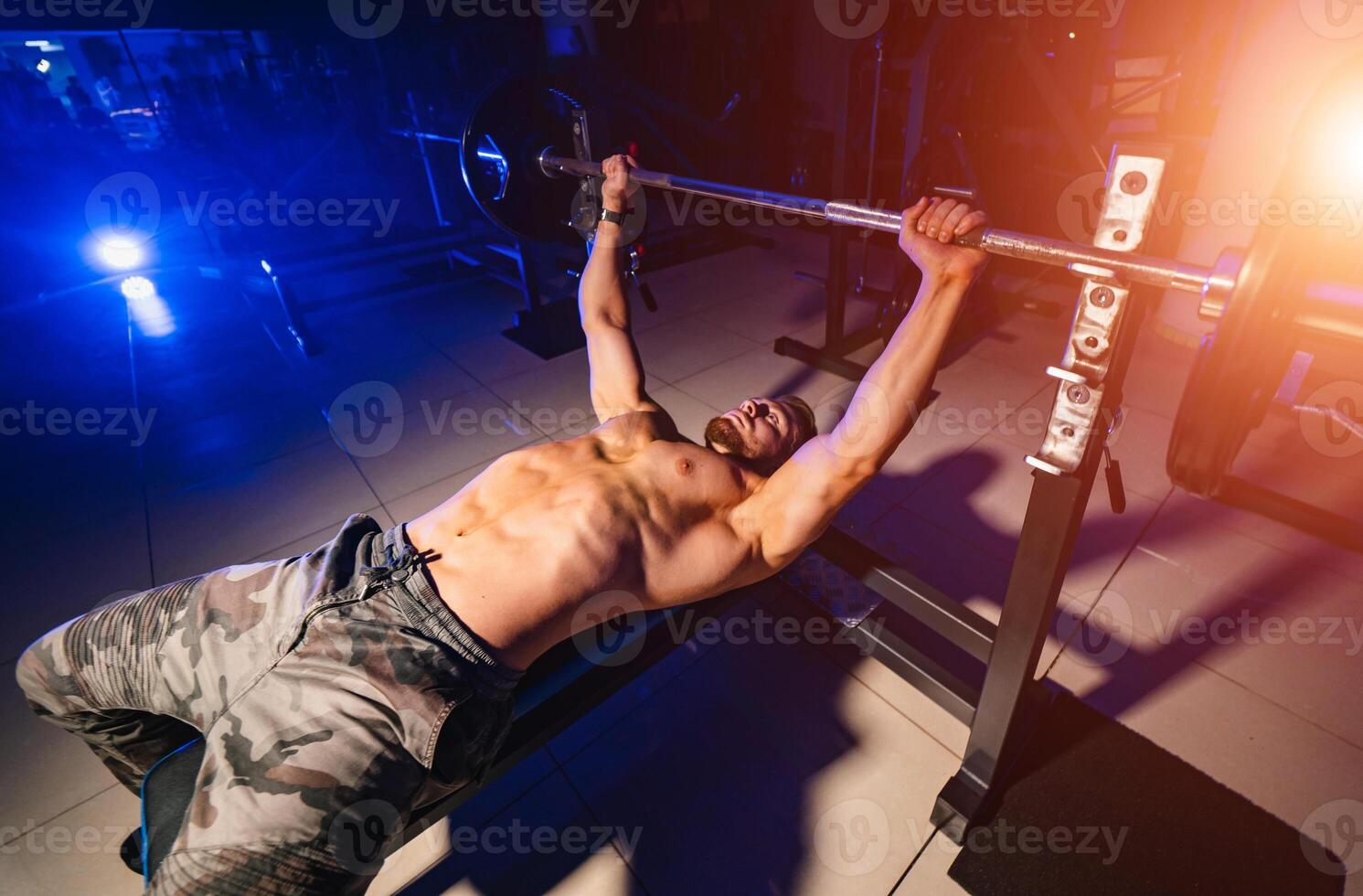 Handsome young man doing bench press workout in gym. Blue light filter. Strong abs. Heavy lift. View from above. Closeup. photo