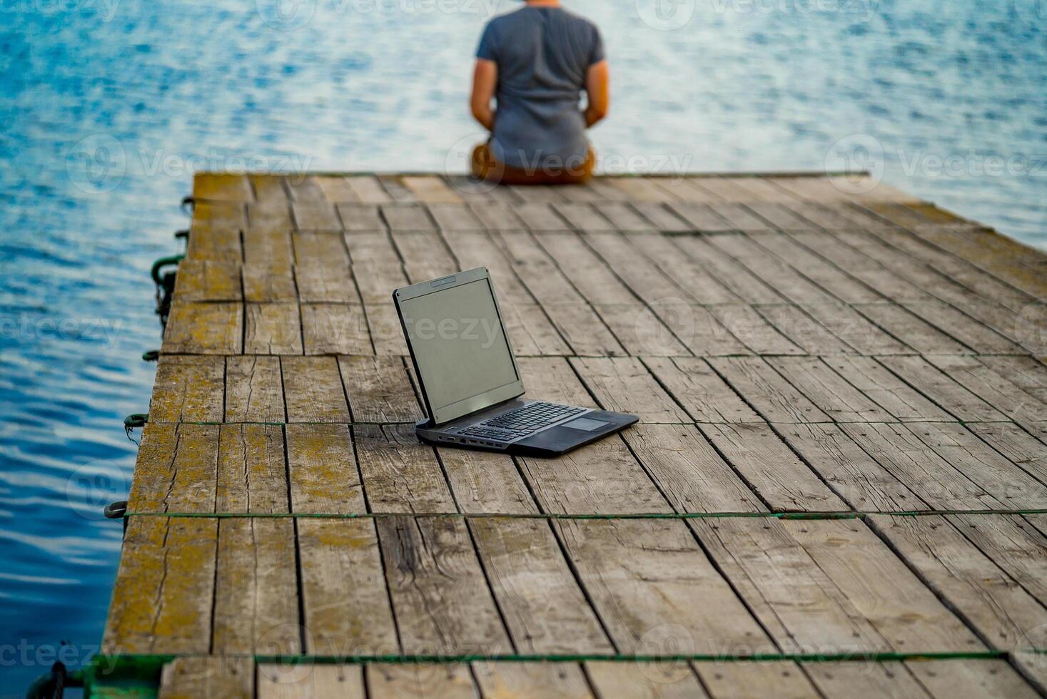 joven hombre sentado en un muelle, utilizando su ordenador portátil. trabajo y vacaciones foto