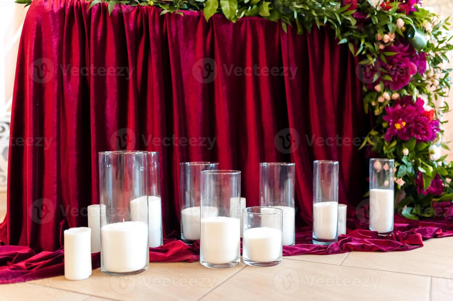 Many candles in glass are standing on the floor on the burgundy background. Romantic design for wedding ceremony decorated with burgundy cloth and flowers with white candles. photo