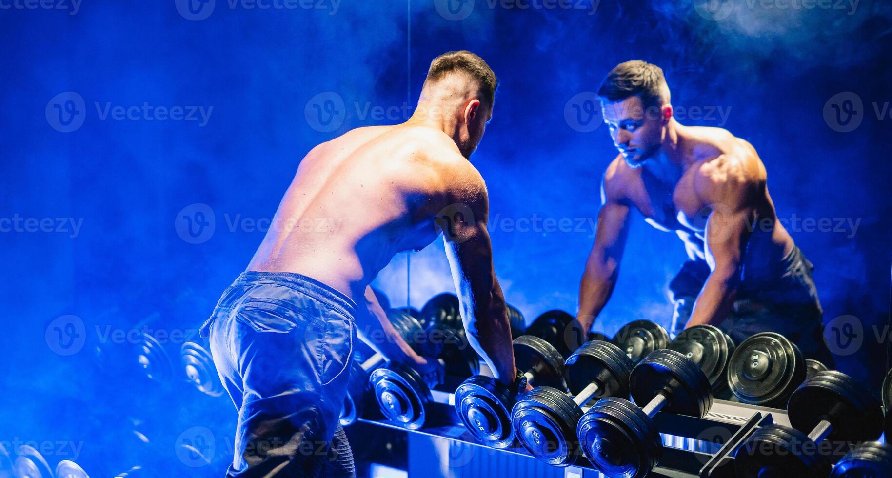 An athlete naked torso man standing in front of a mirror and exercises with dumbbells in the gym, lifestyle health concept. Blue light filter, photo