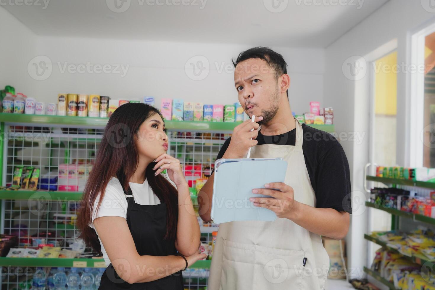 Thoughtful Asian people holding a tablet, cashier is wearing black and cream apron standing in a groceries or convenient store photo