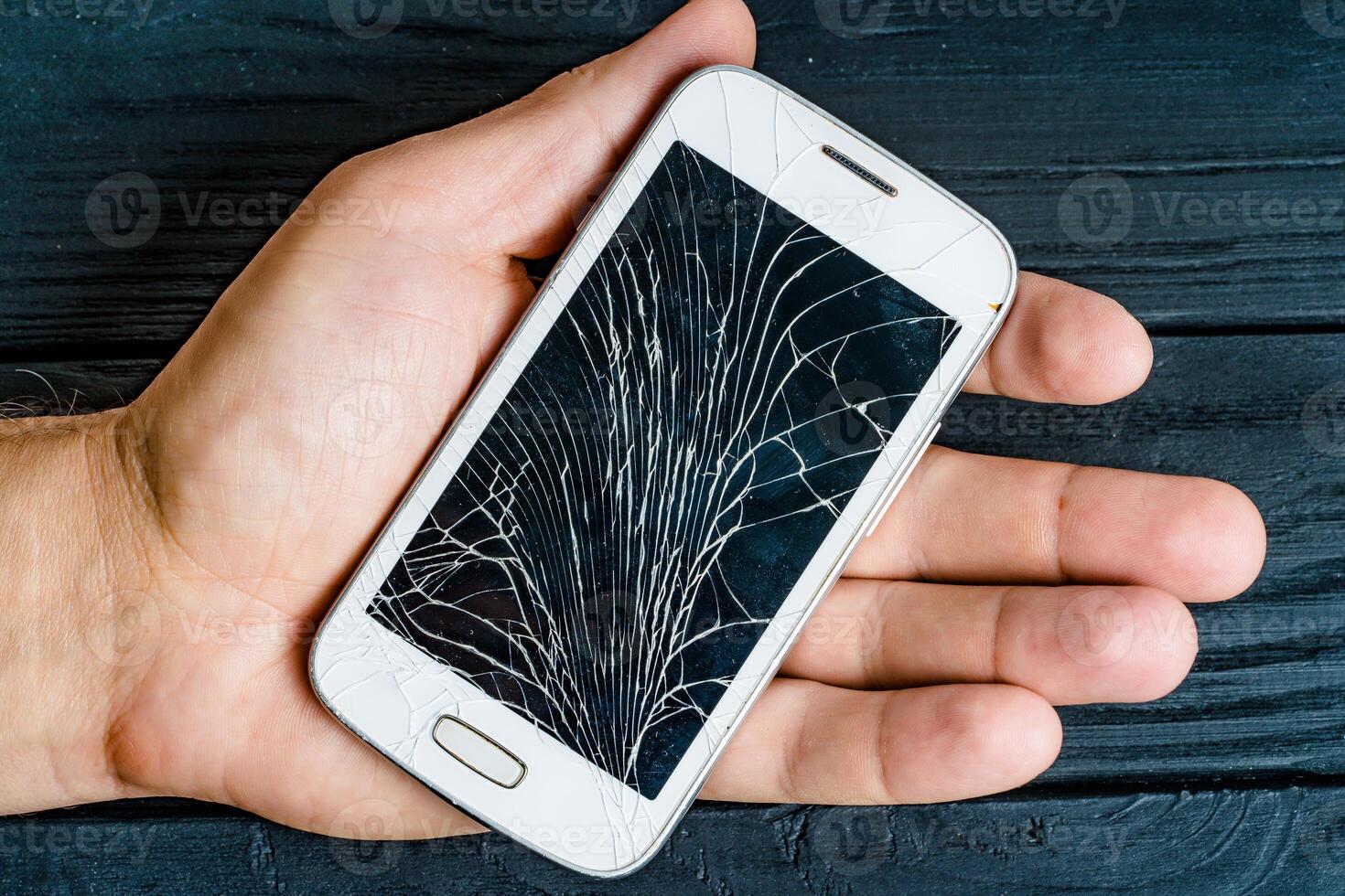 Hand of a male holding white smartphone with damaged glass screen indoors. Broken sensory gadget in the man's hand on the black wooden background. Close-up photo