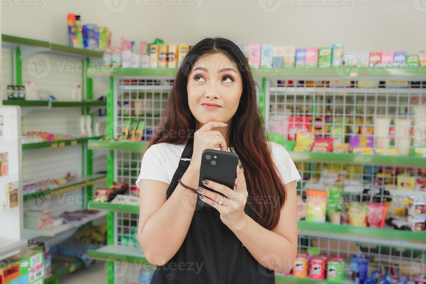 Thoughtful Asian people holding her phone, cashier is wearing black apron standing in a groceries photo