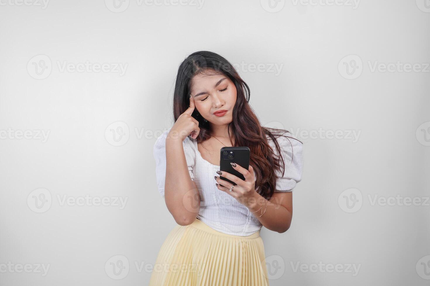 A thoughtful Asian woman is holding her smartphone while imagining her thoughts, isolated by white background. photo