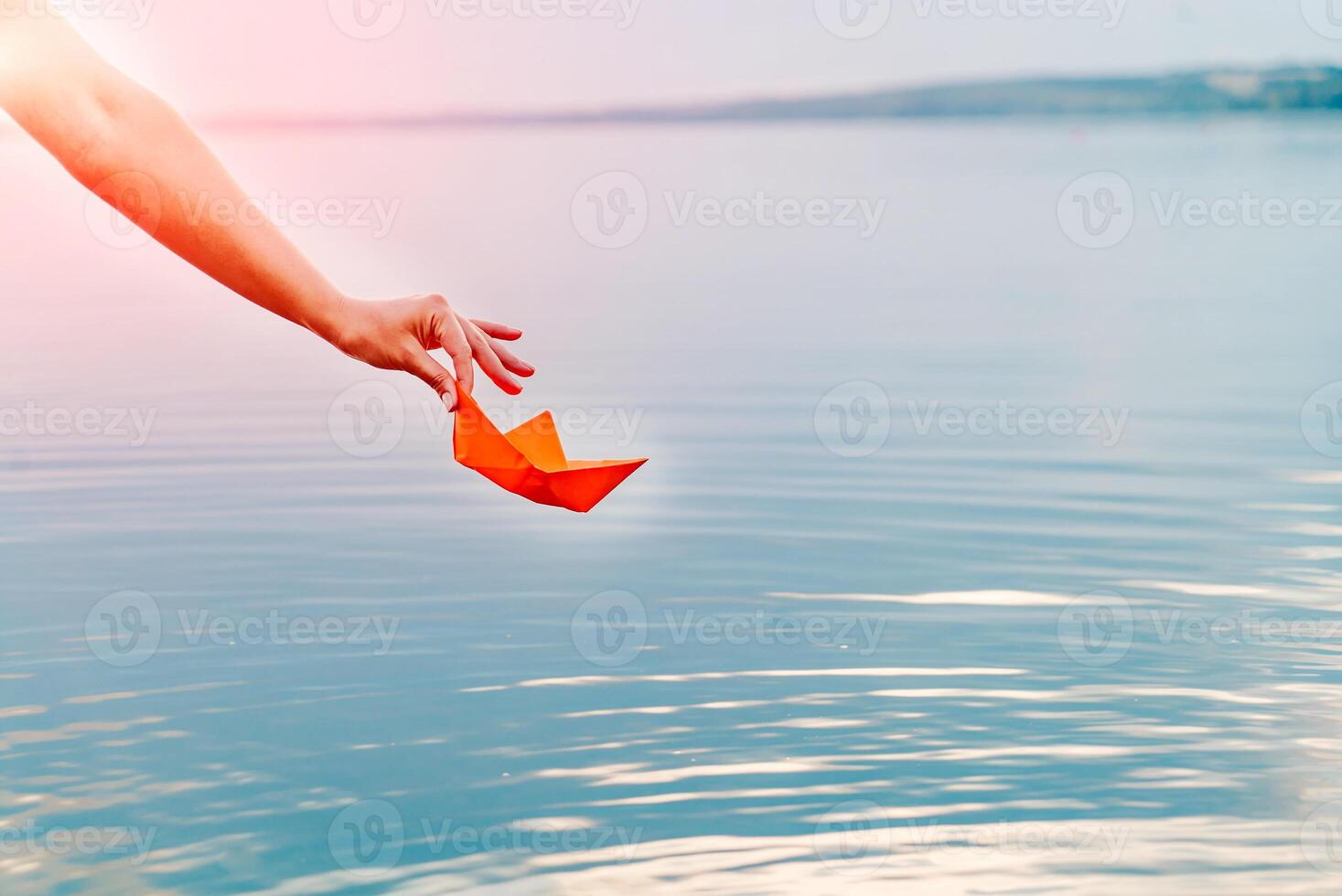 Hands of a young female with a paper boat by the lake in summertime. Woman putting origami boat on water surface. photo