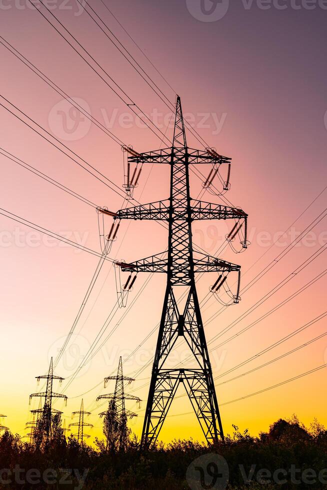 Huge transmission tower with some other towers at the background. Power lines at the scenery of violet sky. Pink sundown with dark tower silhouettes. photo