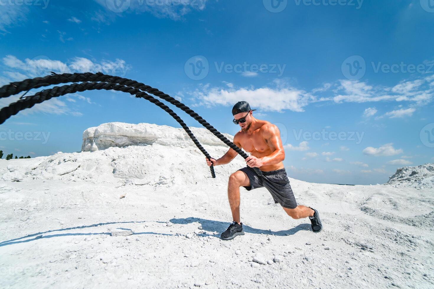 Strong bodybuilder in a cap and sunglasses training with ropes outdoor. Heavy training in a quarry. White landscape. photo