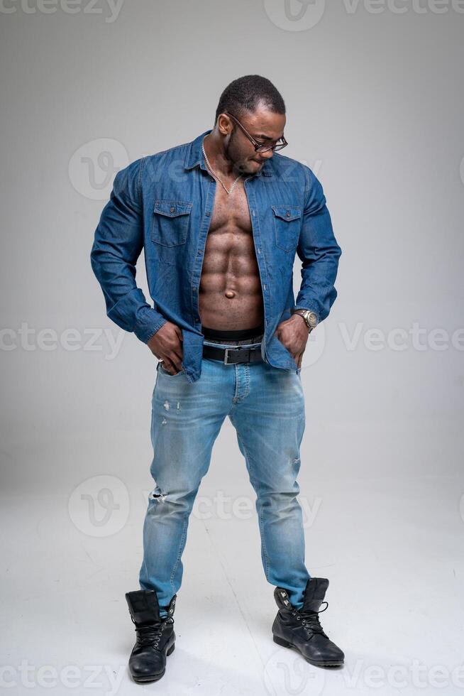 Handsome African guy with naked torso wearing jeans shirt. Athletic man in denim sanding on white background. Bodybuilder male model wearing glasses. photo