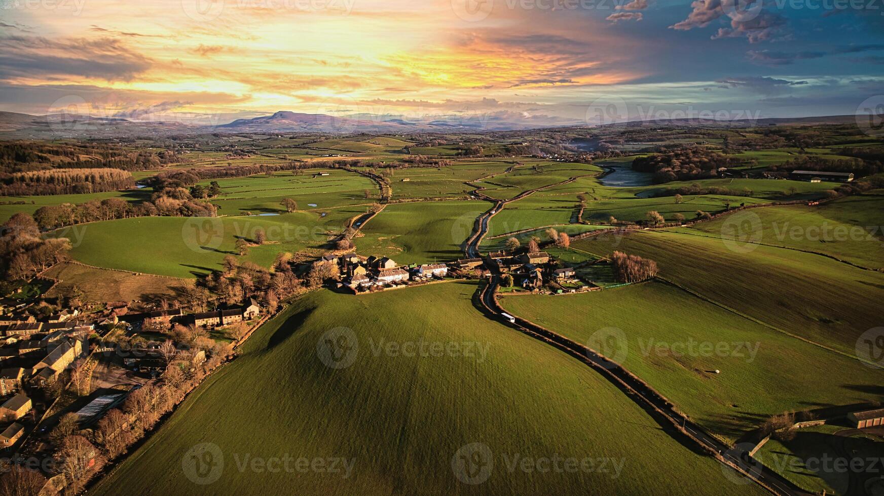Aerial view of a scenic countryside at sunset with lush green fields, a small village, and a winding road leading towards distant hills. photo