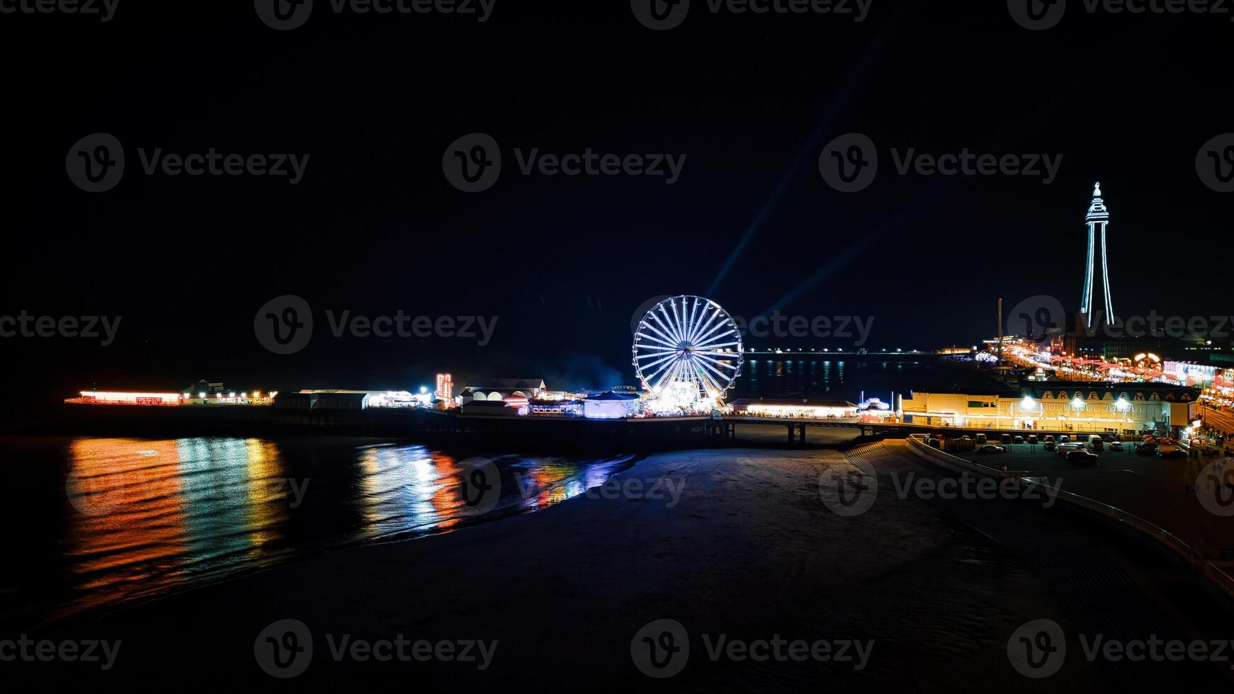 Coastal night scene with illuminated Ferris wheel and lighthouse, vibrant lights reflecting on water, and a dark beach foreground in Backpool, England. photo