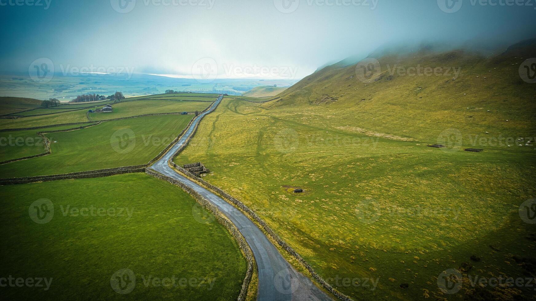 Aerial view of a winding road through lush green hills with mist and a glimpse of the ocean in the distance. photo