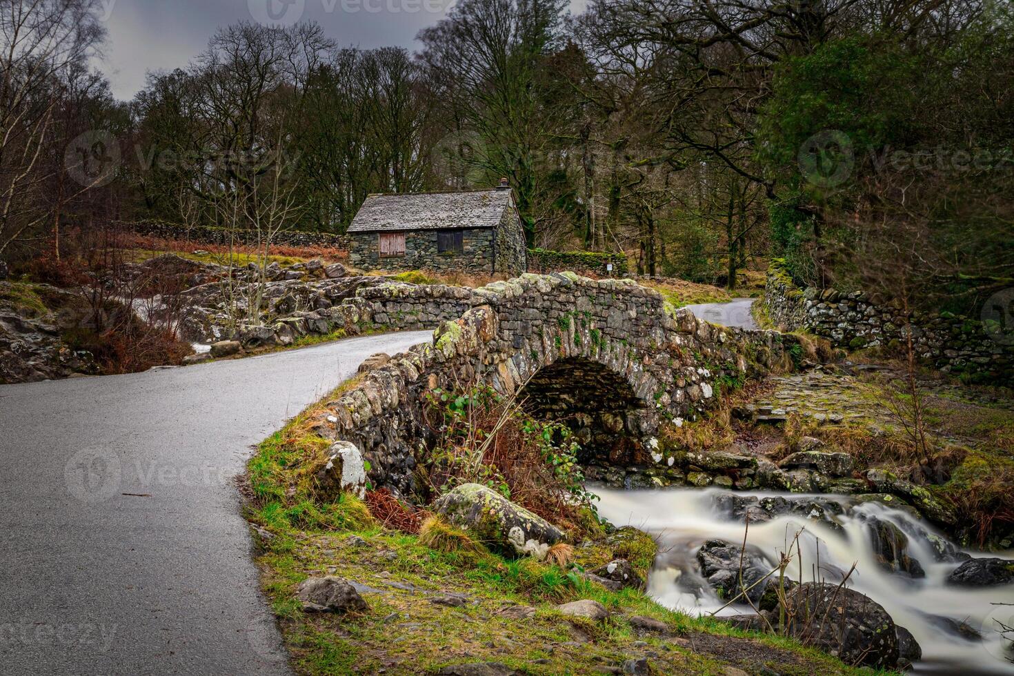 Stone bridge over a stream with a cottage in the background, surrounded by a lush landscape in Lake District. photo