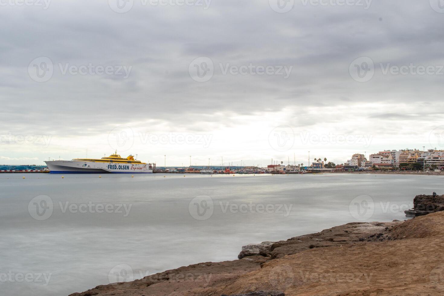 Serene seascape with a ferry boat on the horizon under a cloudy sky, coastal cityscape in the background in Los Cristianos, Tenerife. photo