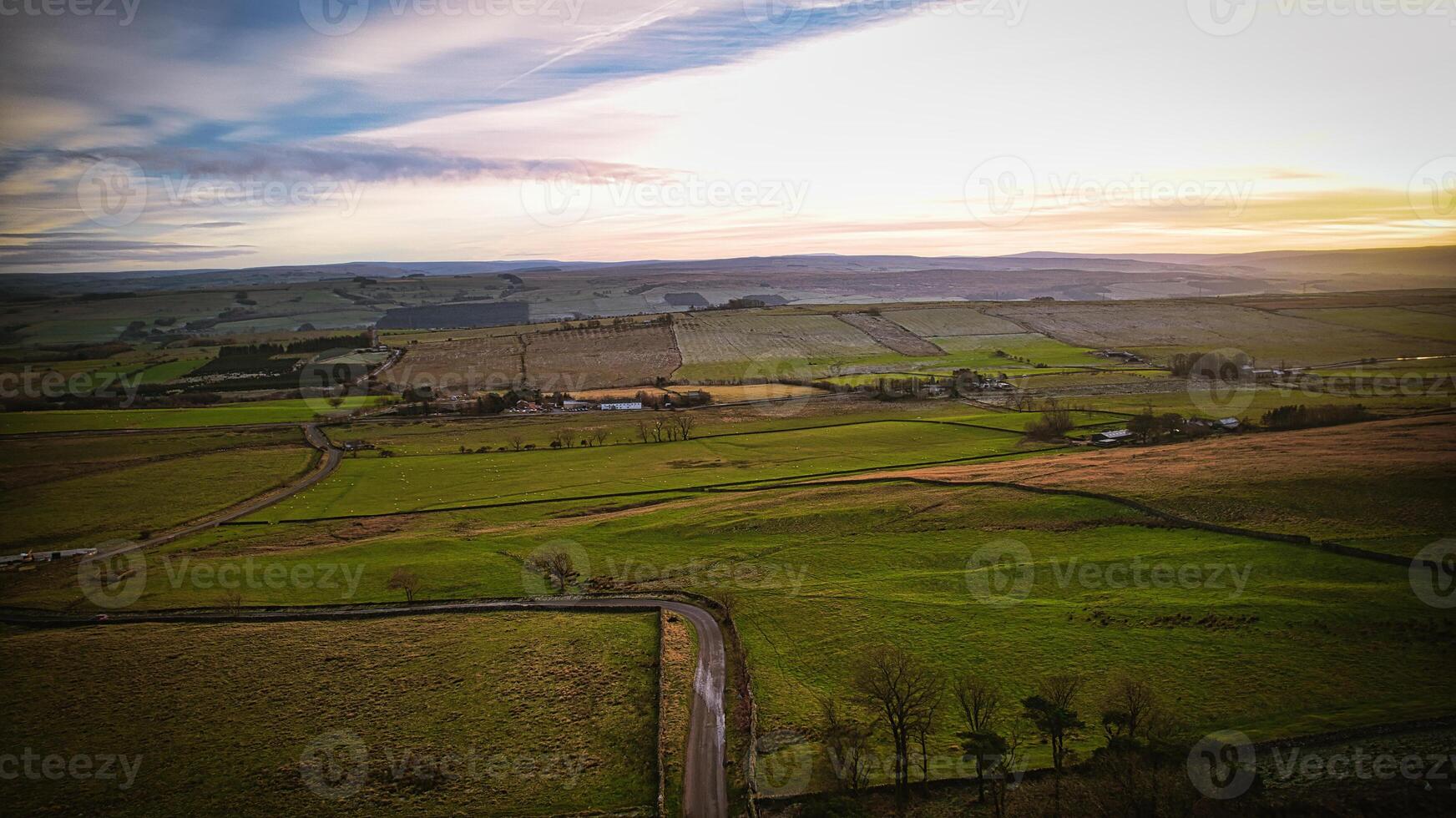 Aerial view of a countryside road at sunset with lush green fields and a soft, colorful sky at Sycamore Gap, Northumberland, UK. photo