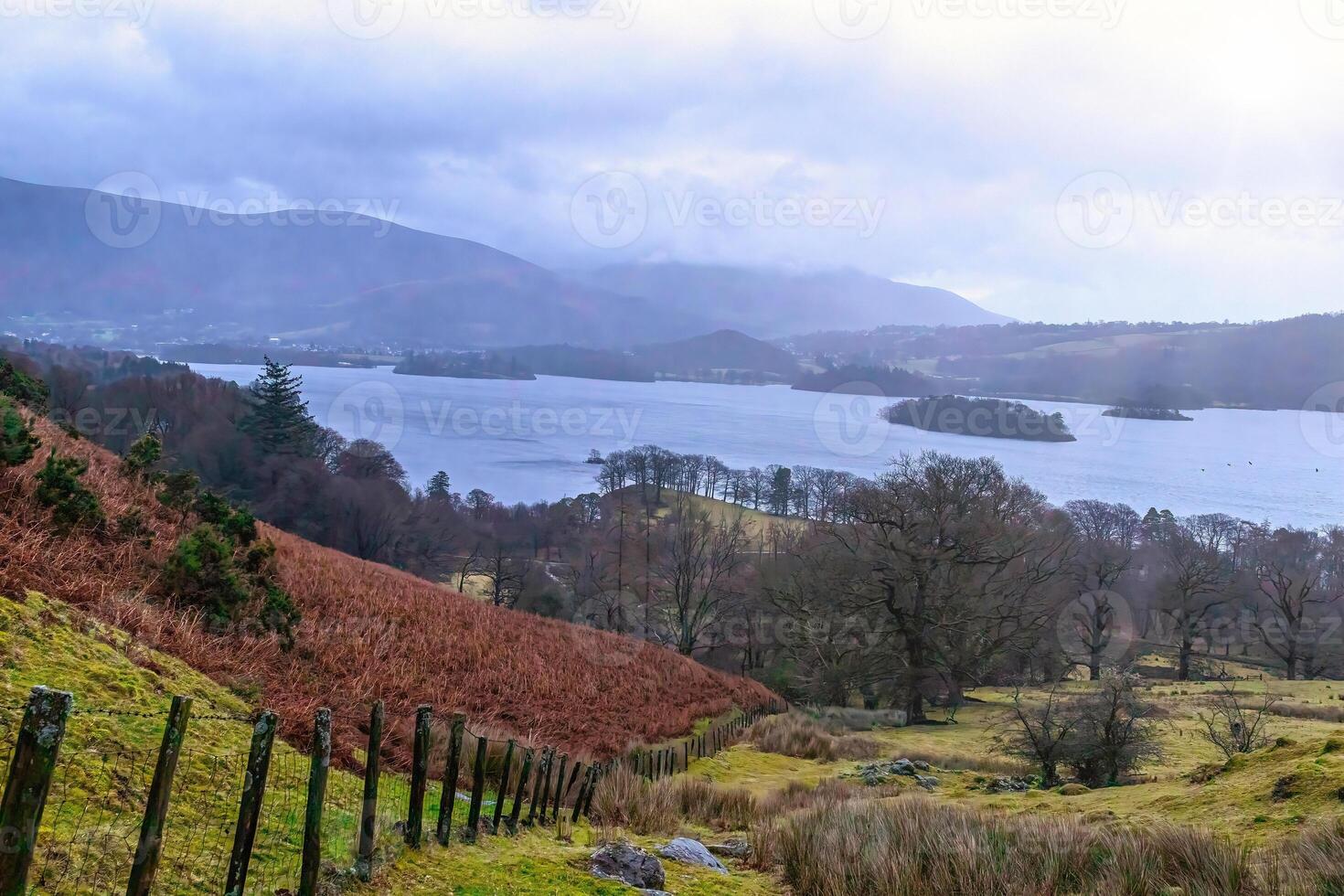 Scenic view of a lush valley with a lake, rolling hills in the background, and a fence leading into the distance on a cloudy day. photo