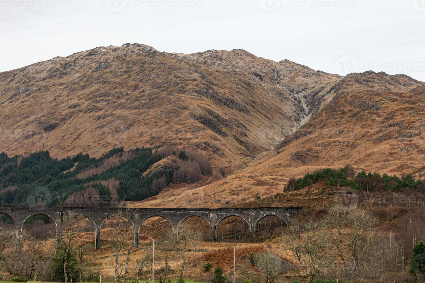 Scenic view of an old viaduct in front of rolling hills and mountains with dry grass and sparse trees in Scotland. photo