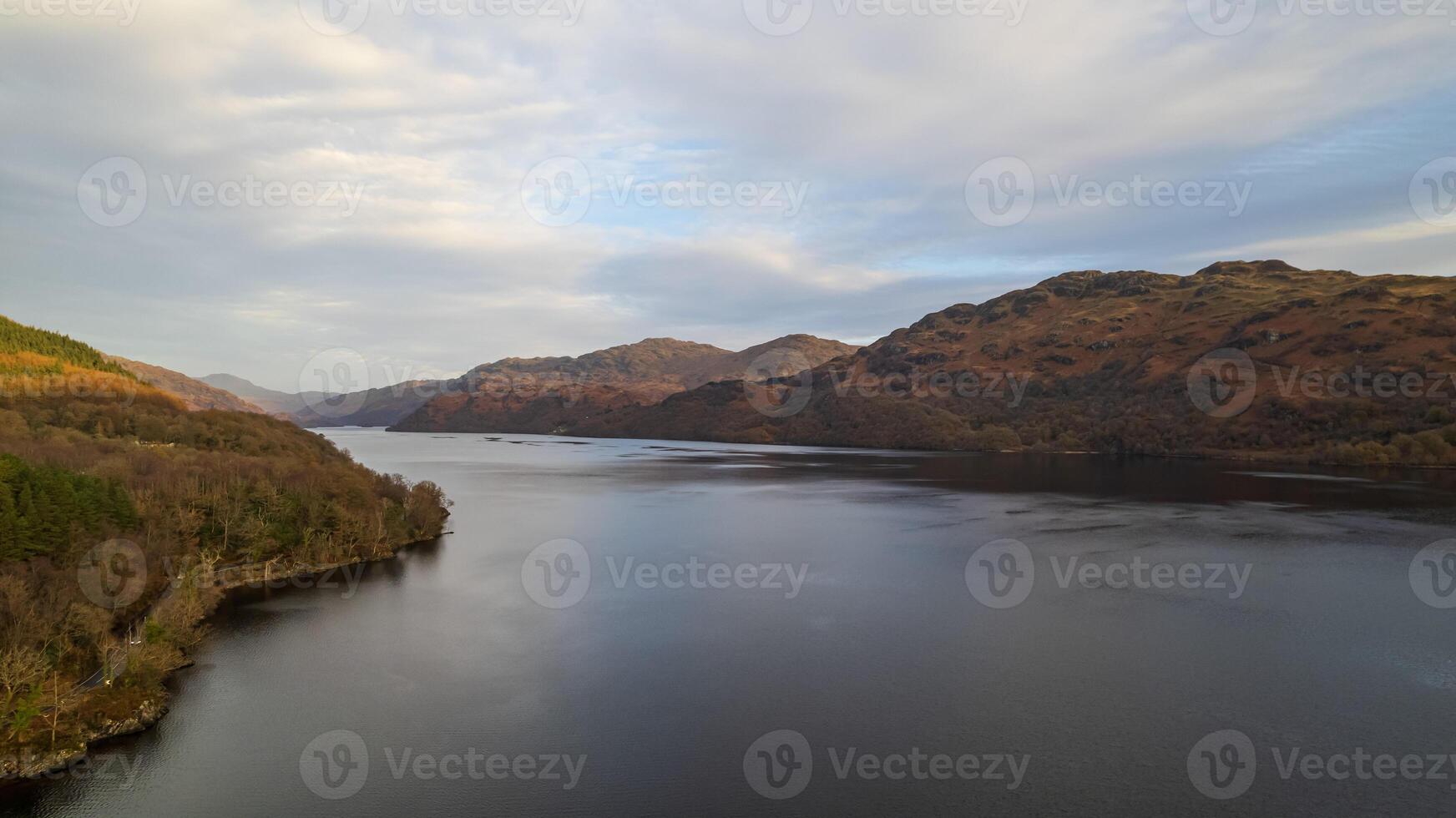 Serene lake with surrounding autumnal hills under a soft cloudy sky, reflecting a tranquil landscape perfect for backgrounds or nature themes. photo