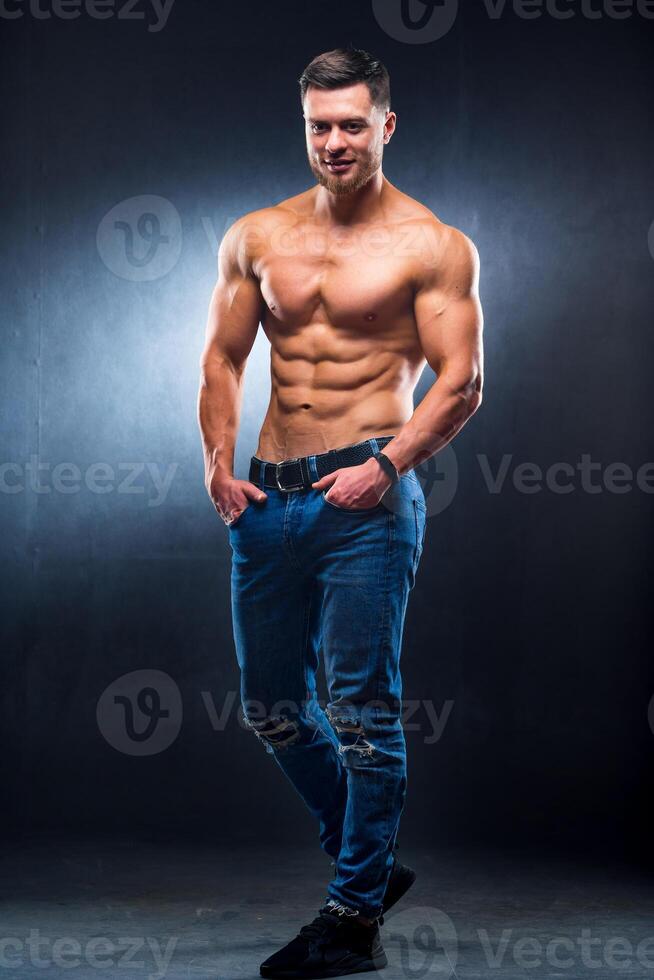 Full size photo of an athletic guy posing with hands in pokets. Studio shot. Bodybuilder. Dark grey background. Closeup.