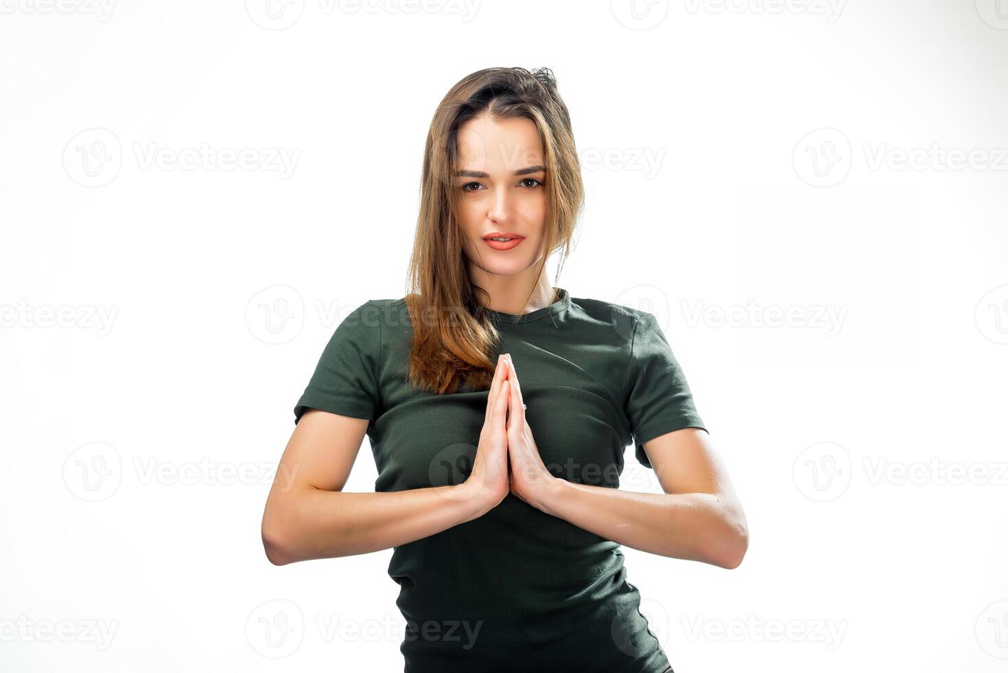 Horizontal shot of positive gorgeous young female with natural make up and long brown hair posing in white studio, pressing hands together in namaste, saying thank you or greeting someone. Closeup photo