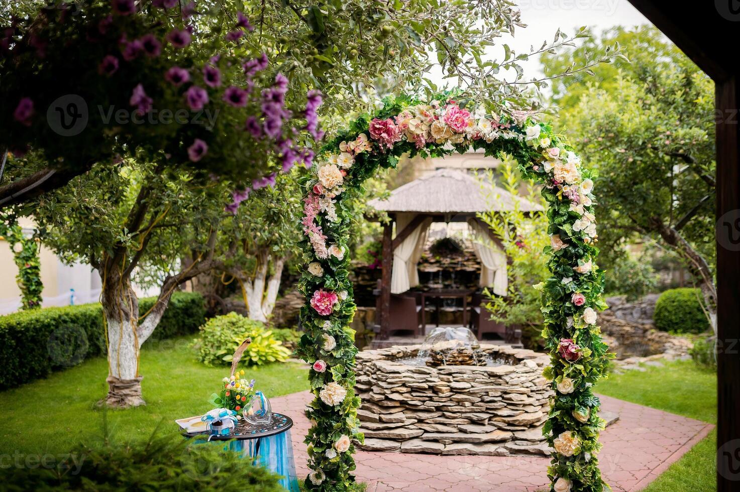 Beautiful arch decorated with colorful flowers on the background of a small fountain outdoors. Natural background in the park with floral archway, green grass and trees in a sunny day. photo