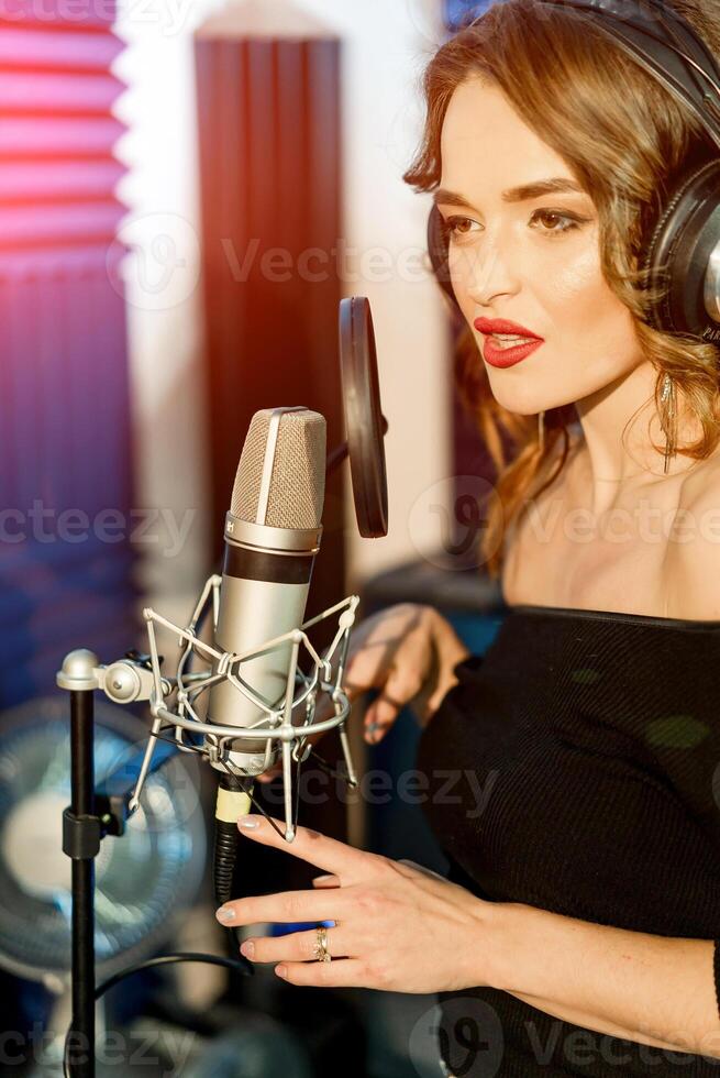 Portrait of a young woman singer with headphones in front of the microphone in the recording studio. Beautiful girl standing near the microphone. photo