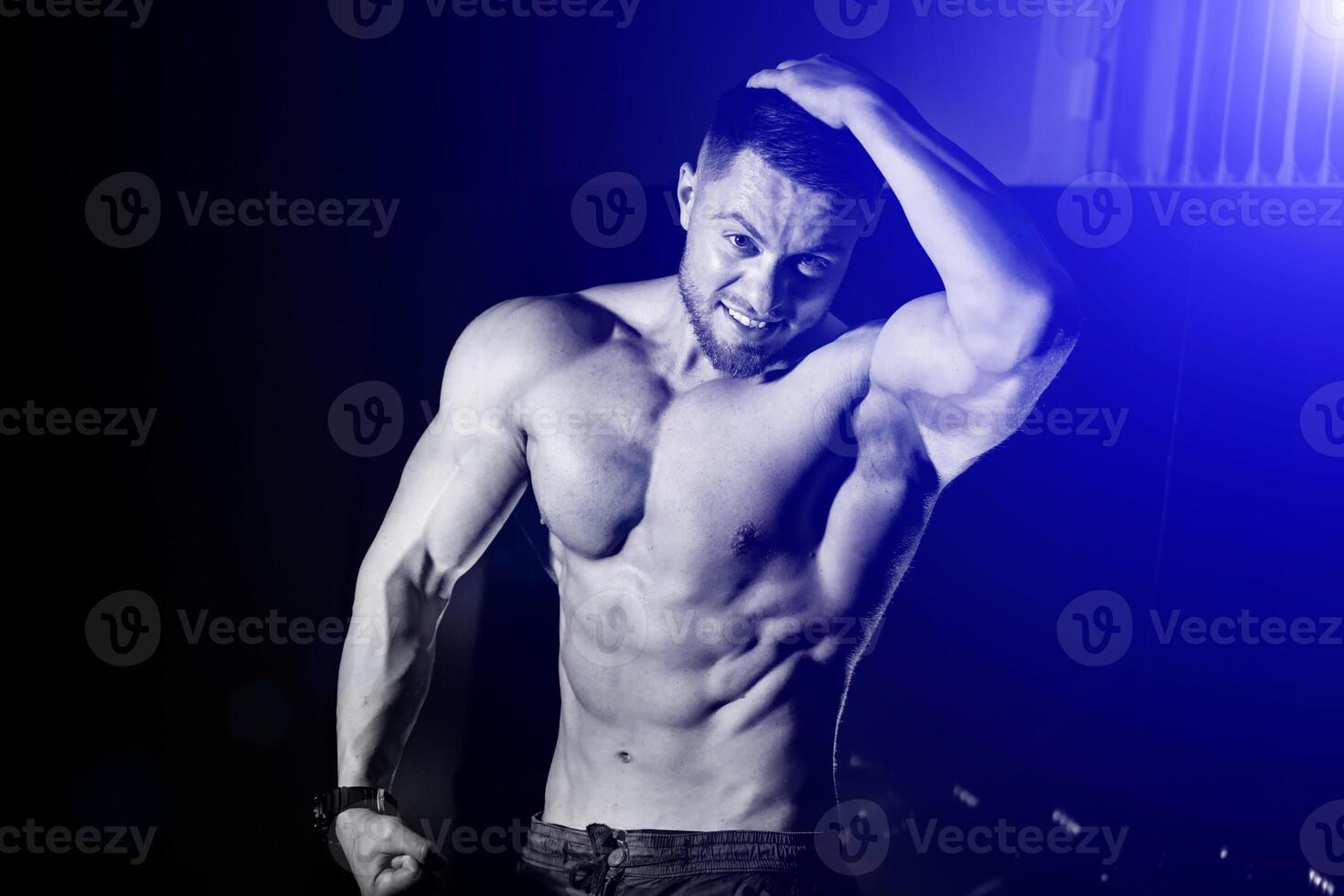 hermoso hombre con grande músculos, posando a el cámara en el gimnasia, negro y azul antecedentes. retrato de un carrocero con desnudo torso. de cerca. foto