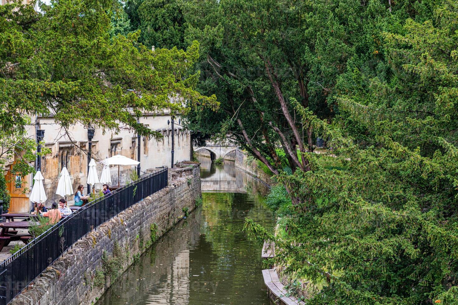 Scenic view of a tranquil canal lined with lush green trees and historic buildings, with outdoor seating by the water's edge in Oxford, England. photo