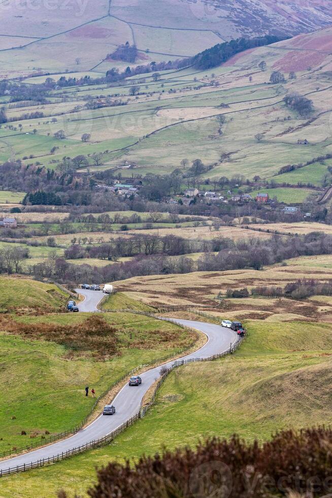 Scenic view of a winding road through a lush valley with rolling hills and a river, under a cloudy sky in Peak District, England. photo
