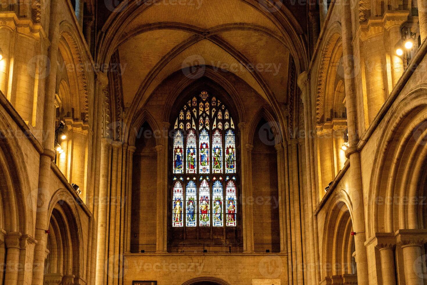 Interior of a Gothic cathedral showcasing an ornate stained glass window with arches and warm ambient lighting in Peterborough, England. photo