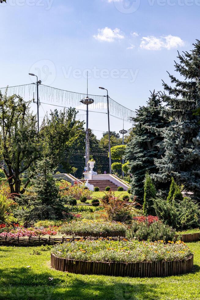 Tranquil park with lush greenery, vibrant flower beds, and a classic fountain under a clear blue sky in Chisinau, Moldova. photo