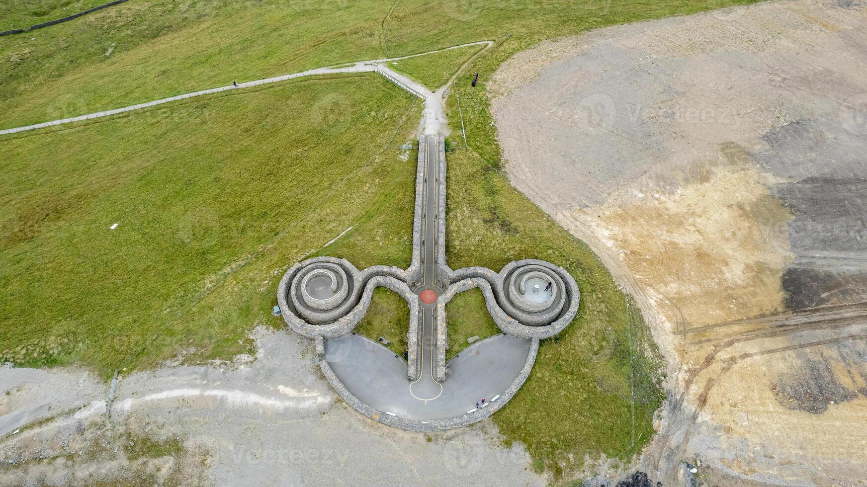 Aerial view of a unique triple-spiral design on the ground, with pathways leading to it amidst a grassy landscape. photo