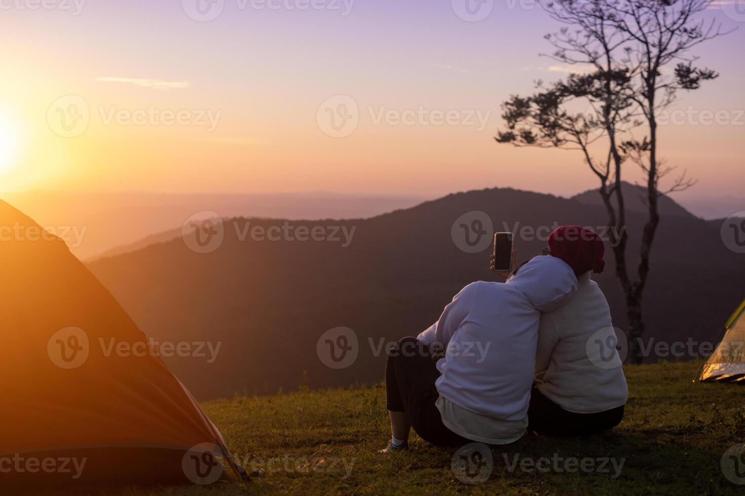 Couple is sitting by the tent during overnight camping and taking selfie while looking at the beautiful scenic sunset over the mountain for outdoor adventure vacation travel photo