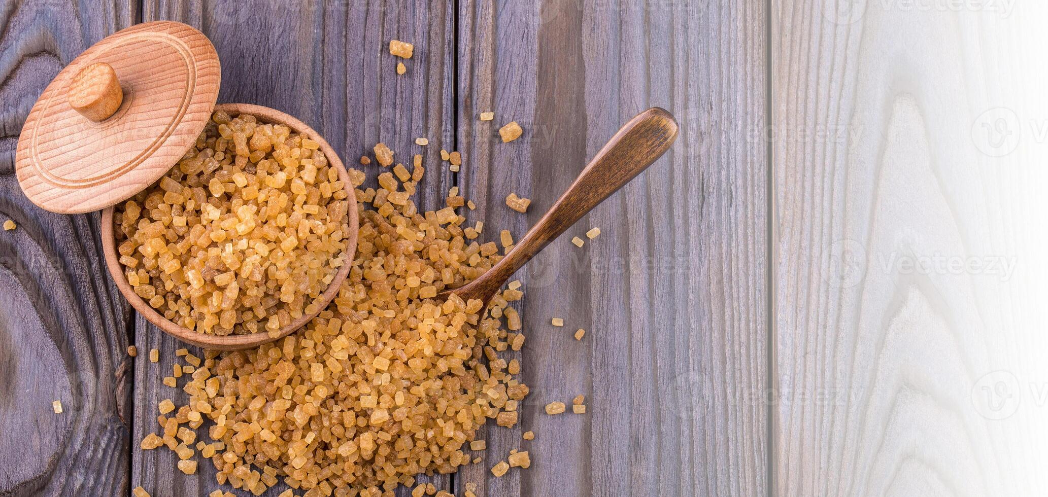 Wooden scoop spoon and bowl full of brown cane sugar with pinch of sugar spilled around on wooden table, top view photo