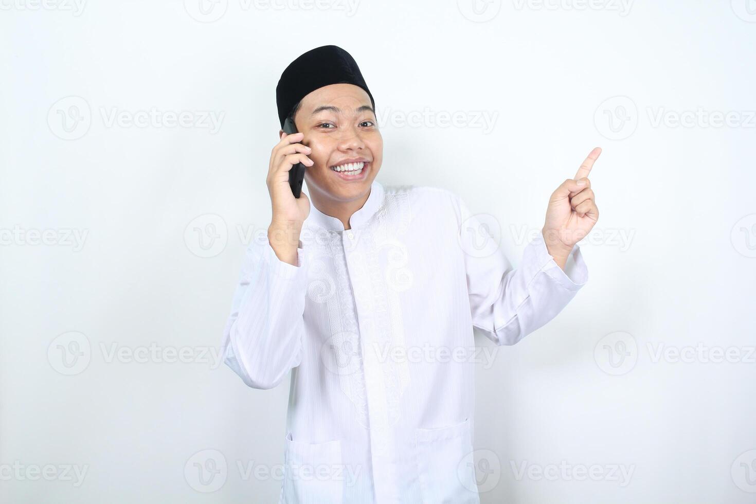 cheerful muslim asian man pointing to beside while speaking on his phone with smiling face isolated photo