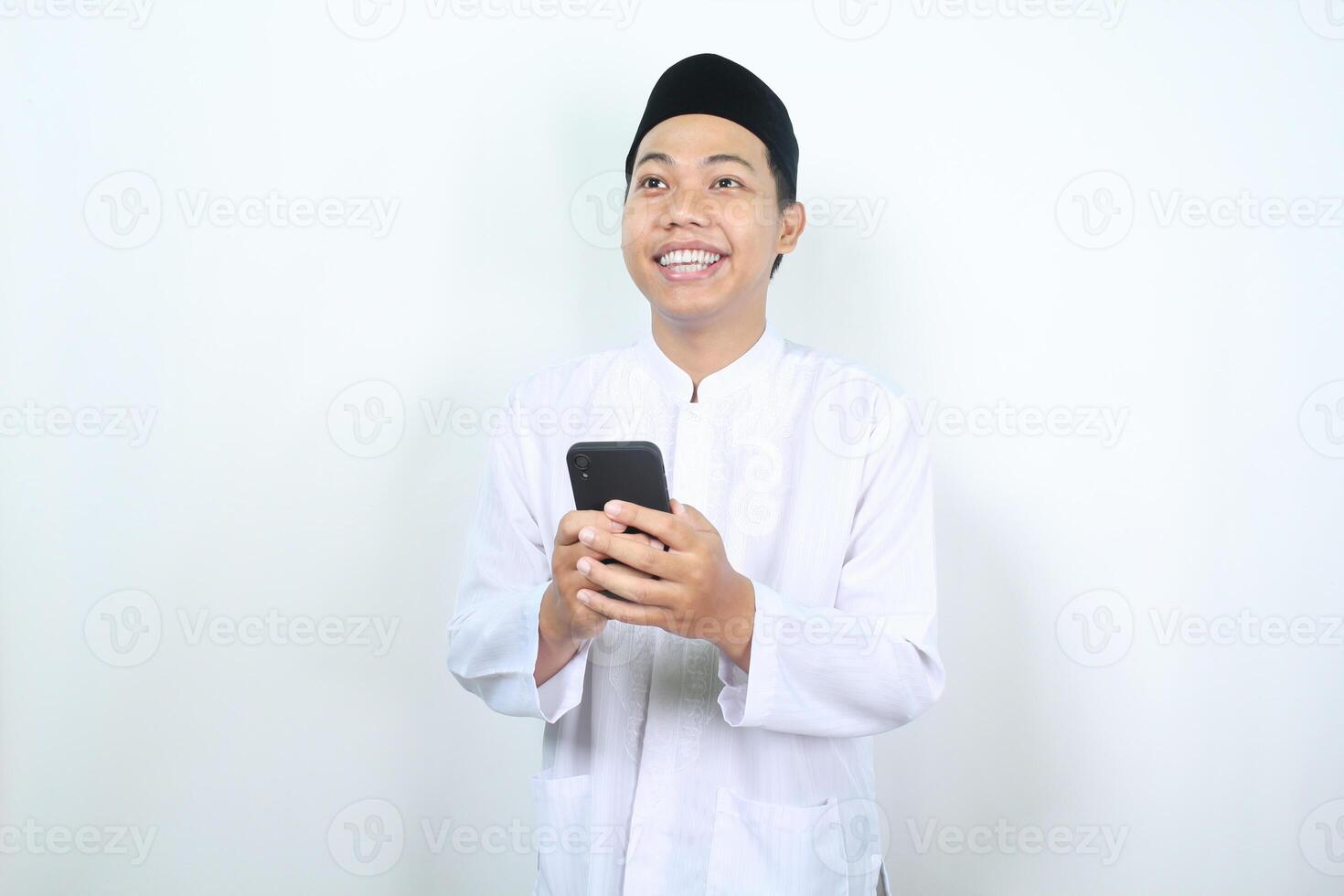 happy asian muslim man smiling while looking above with holding phone isolated photo
