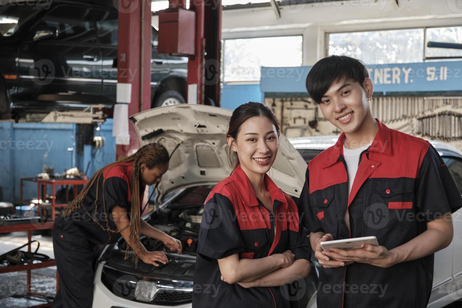 Two Asian professional mechanics, male engineer and partner smile and look at camera, inspect repair work checklist at garage, service car maintenance, and fixing specialist occupations auto industry. photo