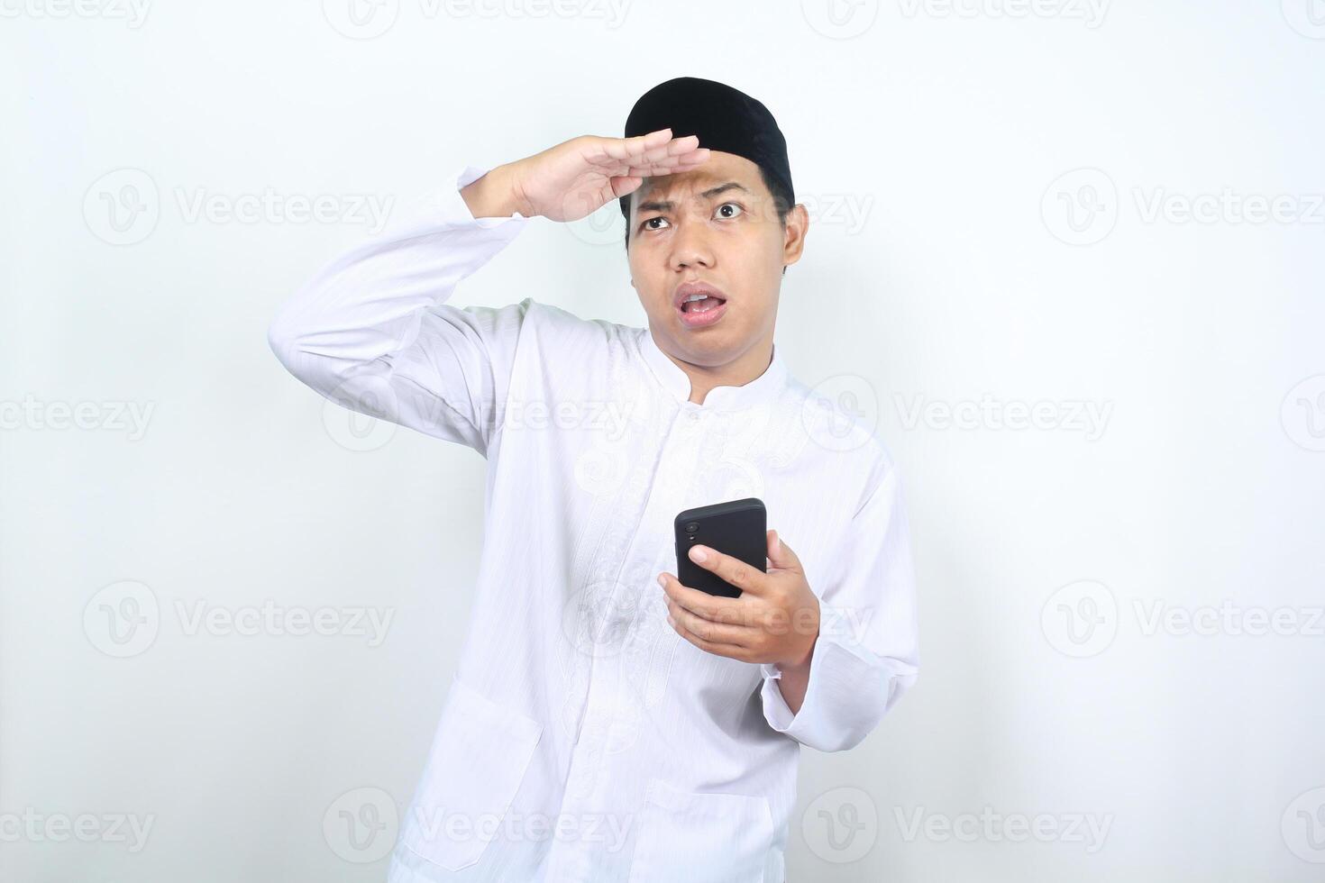 asian muslim man holding mobile phone with raising hand over his head do looking far away gesture isolated on white background photo