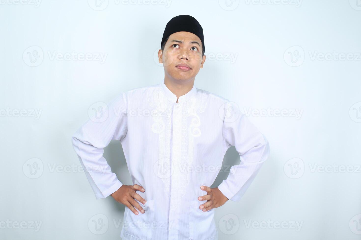 thoughtful asian muslim man looking up with hand on waist thinking something isolated on white background photo