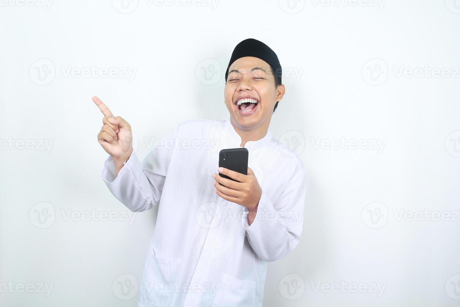 ecstatic asian muslim man laughing while holding phone with pointing finger to the right side isolated photo