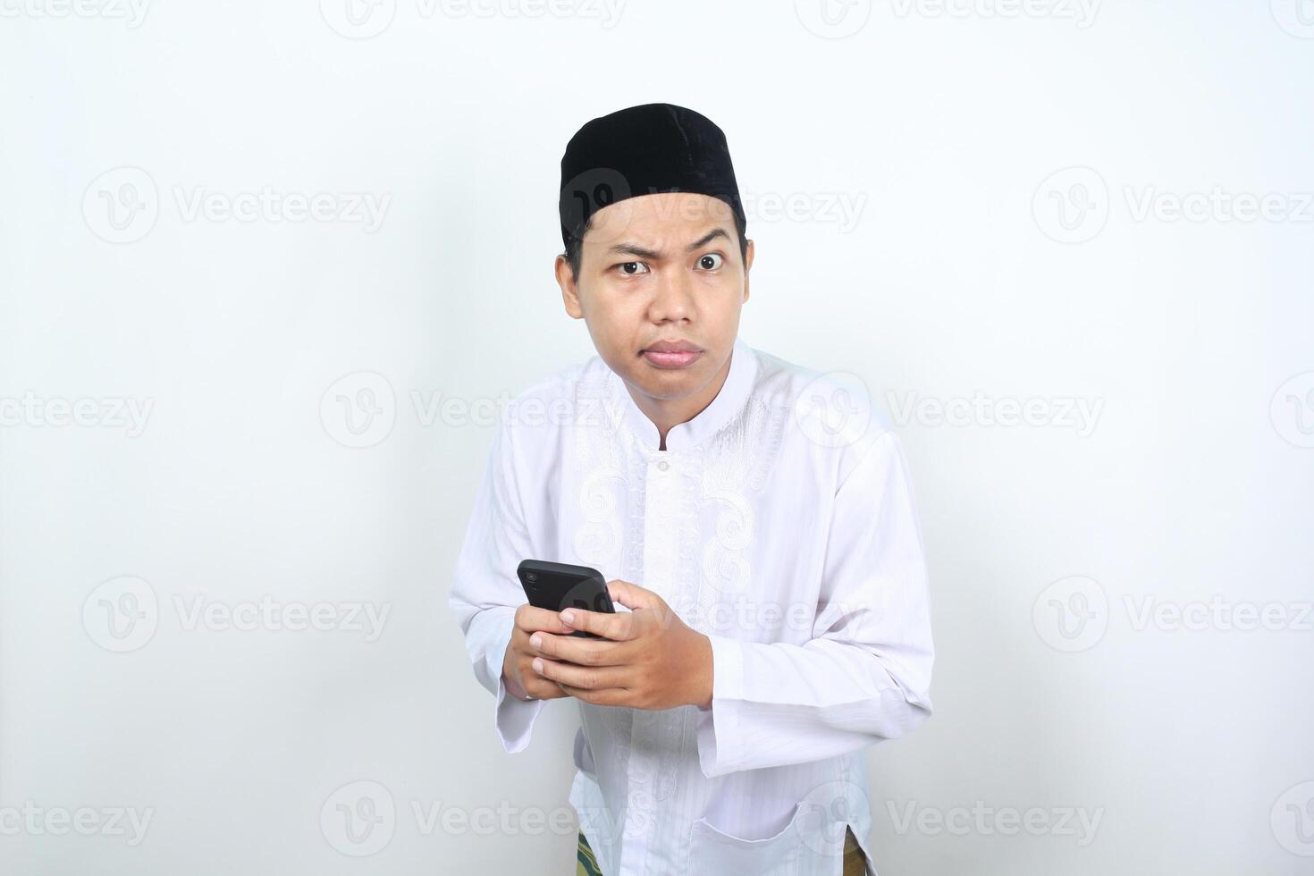 closeup of muslim asian man holding mobile phone looking at camera with confused expression isolated on white background photo