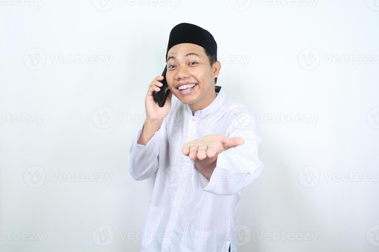 delighted asian muslim man presenting hand forward to camera with smiling face while speaking on his phone isolated photo